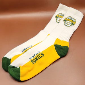 May include: A pair of white athletic socks with the words "SEATTLE SONICS" in green and yellow. The socks have a green heel and toe, and the team logo is printed on the top.