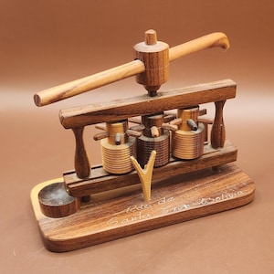 May include: A handcrafted wooden olive oil press model. The miniature press features a lever, three cylindrical containers, and a base with engraved text. The wood has a warm, natural finish, showcasing the grain.