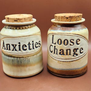 May include: Two ceramic jars with cork lids. One jar reads "Anxieties" and the other reads "Loose Change" in black lettering. The jars are beige with brown and blue accents.