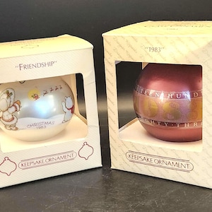 May include: Two Hallmark Keepsake Christmas ornaments in their original boxes. One is white with a winter scene and the word "Friendship". The other is red with "1983" printed on it. Both are in decorative boxes.