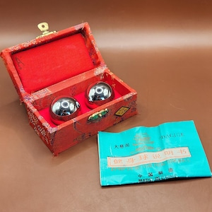 May include: A red, open box with a gold clasp contains two shiny, silver metal balls. The box has a red interior and is lined with red fabric. A teal pamphlet with Chinese text and the word "DACIGE" is next to the box.