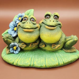 May include: A ceramic frog figurine featuring two smiling frogs sitting on a lily pad. The frogs are green and yellow with large eyes. Blue flowers and green leaves are behind the frogs.