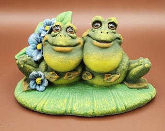 May include: A ceramic frog figurine featuring two smiling frogs sitting on a lily pad. The frogs are green and yellow with large eyes. Blue flowers and green leaves are behind the frogs.