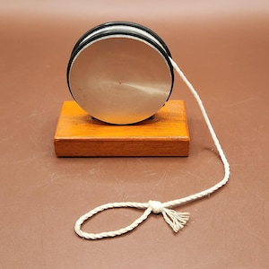 May include: A silver yo-yo with black rubber rings, resting on a small, rectangular wooden base. The yo-yo has a white string attached, knotted at the end. The wooden base is stained a warm orange color.