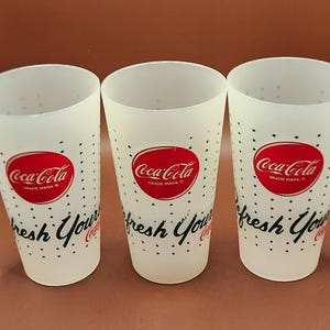 May include: Three white Coca-Cola drinking glasses. Each glass has a red Coca-Cola logo with the words "Trade Mark" and the phrase "fresh Your" in black script. The glasses are decorated with small black polka dots.