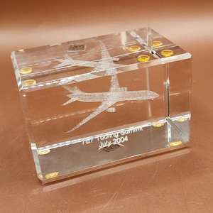 May include: A clear glass cube with etched airplane designs. The cube has gold-colored accents and text that reads "7E7 Tooling Summit July 2004". The cube is a commemorative item.