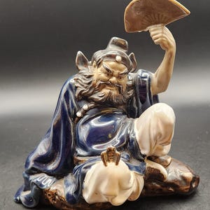 May include: A ceramic figurine of a seated, bearded man in a blue robe, holding a fan. The figurine has a detailed face and is resting on a brown base. The fan is a light brown colour.