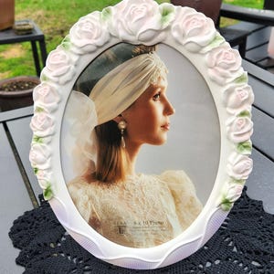 May include: Oval white picture frame with a floral rose design. The frame holds an 8 x 10 photo of a woman in a wedding dress. The frame has a scalloped edge and is sitting on a black lace doily.