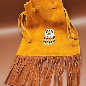 May include: Mustard yellow suede pouch with a beaded owl design. The pouch has a drawstring closure and a fringe bottom. The owl design features white, black, and blue beads. The fringe and drawstring are a darker brown.