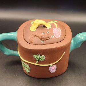 May include: A brown ceramic teapot with a square body and a lid featuring a yellow accent. The handle and spout are turquoise. The teapot is decorated with painted leaves and berries.