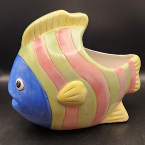May include: A ceramic fish-shaped planter. The head is blue, the fins are yellow, and the body is striped with pink and green. It has a wide opening for plants and a whimsical design.