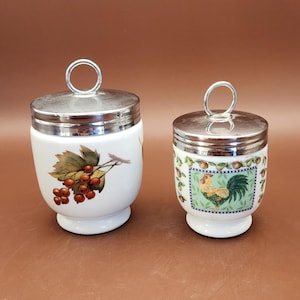 Puede incluir: Dos hueveras de cerámica blanca con tapas y asas plateadas. Una taza presenta un diseño de bayas rojas y hojas verdes, mientras que la otra tiene una ilustración de gallo dentro de un borde decorativo. Las hueveras están sobre una superficie marrón.