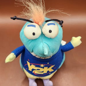 Puede incluir: Un peluche con forma de personaje con cuerpo azul claro, ojos blancos grandes y un mechón de pelo naranja y marrón claro. Lleva una camiseta azul con el texto "V2K" en amarillo. El juguete tiene pequeñas alas y pies morados.