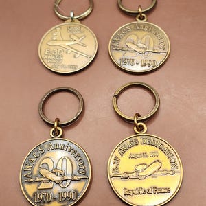 May include: Four bronze-colored keychains with circular pendants. Each pendant features an airplane design and text. One reads "E-3D AWACS Rollout July 11, 1989 United Kingdom." Others say "AWACS Anniversary 1970-1990" and "E-3F AWACS Dedication August 22, 1990 Republic of France."