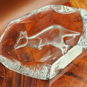Op de afbeelding: Een presse-papier van helder glas met een amberkleurig middenstuk met een geëtst ontwerp van een vos in profiel. Het glas heeft een gladde, onregelmatige, ronde vorm met getextureerde, matte details langs de onderrand van het object.