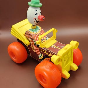 May include: Vintage Fisher-Price Jalopy toy car featuring a clown driver. The car has a yellow body, orange wheels, and a brown base. The clown has a white head, red nose, and green hat. The text "JALOP" is visible on the car.