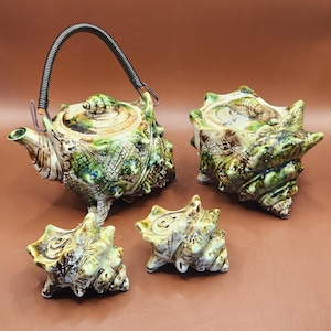 May include: A ceramic teapot and sugar bowl set with a seashell design. The teapot has a black handle and spout. The set includes a teapot, a sugar bowl with a lid, and two small seashell-shaped pieces. The colors are green, brown, and cream.