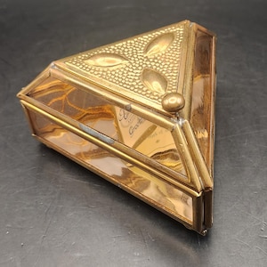 May include: A vintage, triangular-shaped trinket box with a gold-colored metal frame and clear glass sides. The top is decorated with embossed floral designs and a textured pattern. The box contains a card with the word "Greetings" visible.