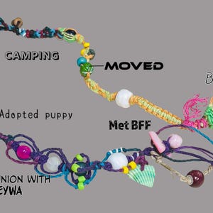 May include: A handcrafted macrame necklace with various beads, shells, and decorative elements. The necklace features text labels including "Camping", "Adopted puppy", "Moved", "Met BFF", "Birth", and "Communion with EYWA".