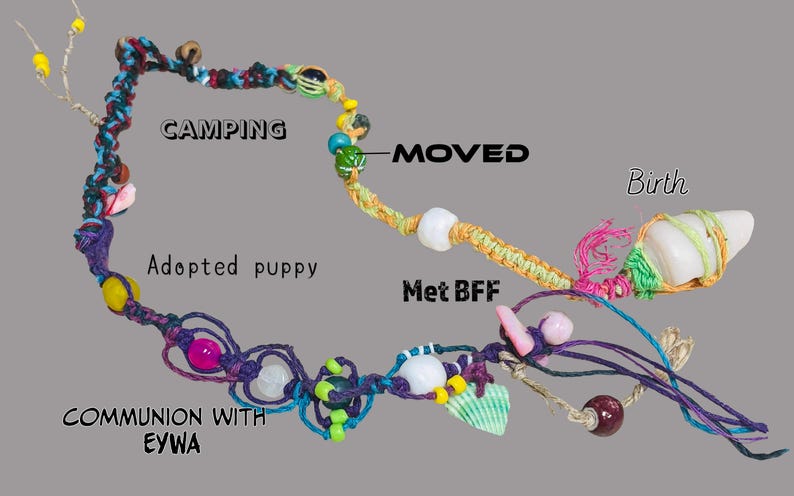 May include: A handcrafted macrame necklace with various beads, shells, and decorative elements. The necklace features text labels including "Camping", "Adopted puppy", "Moved", "Met BFF", "Birth", and "Communion with EYWA".