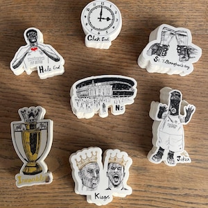Op de afbeelding: Een verzameling witte stickers met zwarte en gouden illustraties. De stickers tonen een klok, een stadion, een trofee en portretten van twee mensen met kronen. Andere stickers tonen figuren met tekst, waaronder "Hale End", "St. Totteringhon's day", "Invincible", "Kings" en "Joker".