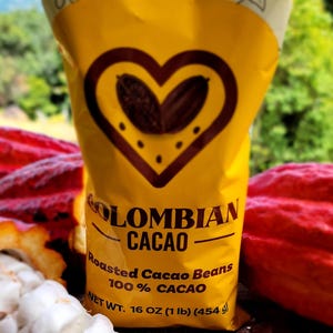 May include: A yellow bag of Colombian Cacao with a heart-shaped design containing cacao beans. The bag reads "Roasted Cacao Beans 100% Cacao" and "NET WT. 16 OZ (1 lb) (454 g)". Cacao beans and pods are visible.