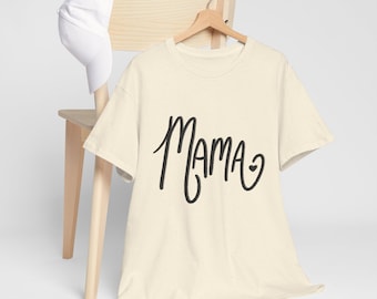 May include: Cream-colored t-shirt with the word "Mama" in black script, featuring a small heart. The shirt hangs on a wooden hanger, with a white baseball cap resting on a wooden chair. The shirt has a speckled texture.