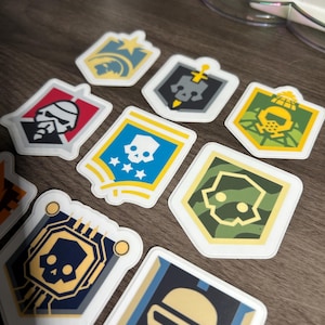 Puede incluir: Una colección de pegatinas coloridas, troqueladas, con varios diseños de escudos. Las pegatinas presentan imágenes de calaveras, estrellas y otros elementos gráficos en tonos azules, amarillos, rojos y verdes. Las pegatinas están sobre una superficie de madera.