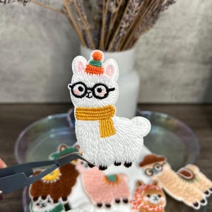 May include: Embroidered llama patches in various colors, including white, brown, and pink. The white llama wears glasses, a hat, and a scarf. Other patches feature similar designs. The patches are on a clear plate.