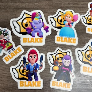 May include: A collection of colorful character stickers featuring various designs. Each sticker has a white border and the name "BLAKE" printed on a yellow background. The stickers depict cartoon figures with unique outfits and accessories.