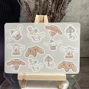 Puede incluir: Una hoja de pegatinas con personajes de dibujos animados en varias poses. Las pegatinas son principalmente blancas y marrones, algunas llevan sombreros, sujetan globos o llevan paraguas. Las pegatinas se muestran en un pequeño caballete de madera.