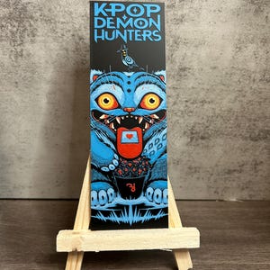 Può includere: Una stampa rettangolare nera con la scritta "KPOP DEMON HUNTERS" in blu. L'immagine presenta un mostro blu con occhi gialli, che tiene in mano una tazza con bacche rosse. Il mostro ha un cuore sulla lingua.