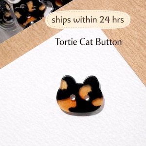 May include: A Tortie Cat Button with a black base and orange and tan spots. The button is shaped like a cat's head with two holes for sewing. The text "Tortie Cat Button" is visible.