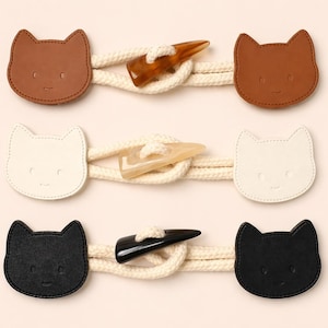 May include: Three cat-shaped toggle closures in brown, white, and black. Each closure features a smiling cat face and is connected by a cream-colored rope with a horn-shaped toggle. The closures are designed for coats, sweaters, or other garments.