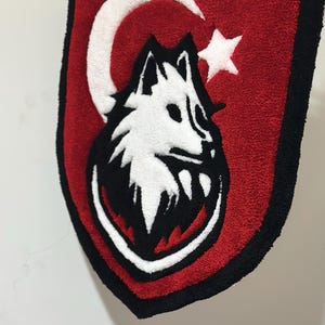 Könnte beinhalten: Ein schildförmiger Teppich mit rotem Hintergrund, schwarzem Rand und einem weißen Halbmond- und Sterndesign. Der Teppich zeigt einen Wolfskopf in Schwarz und Weiß.