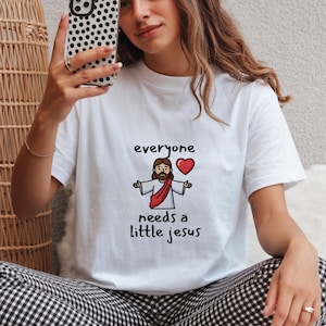 May include: White t-shirt with the text "everyone needs a little jesus" and a graphic of Jesus with a red heart. The shirt is made of a soft material.
