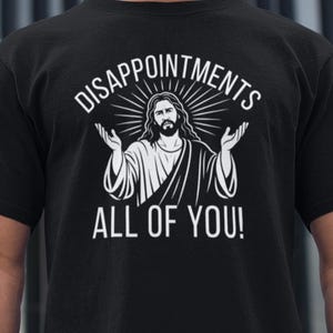 Puede incluir: Camiseta negra con un gráfico blanco de Jesús con los brazos extendidos, rodeado de líneas radiantes. El texto superior dice "DISAPPOINTMENTS" y debajo "ALL OF YOU!"