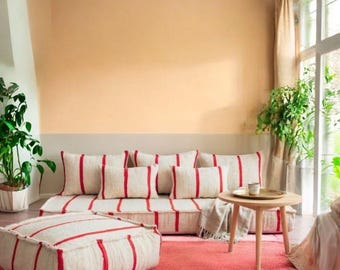 Op de afbeelding: Een woonkamer met een laag profiel bank en bijpassende poef, beide in crème met rode strepen. De kamer bevat een ronde houten bijzettafel, een roze en wit gestreept tapijt en diverse kussens.