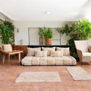 May include: An outdoor seating area with a cream-coloured sofa and cushions, a wooden chair, and a small table. Two cream-coloured rugs sit on the terracotta tiled floor. Green plants surround the space, creating a natural feel.
