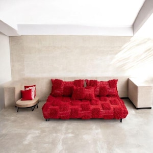 May include: A modern red sofa with matching pillows and a small side chair. The sofa and pillows have a textured, tufted design. The side chair has a beige cushion and a black metal frame. The room has neutral-colored walls and flooring.