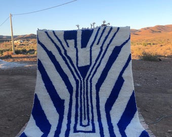 May include: A large, rectangular rug with a bold geometric design in white and blue. The rug features a central pattern of vertical and angled stripes, with a fringe along the bottom edge. The rug is displayed outdoors.