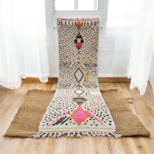 Op de afbeelding: Een crèmekleurig, patroonkleed met een geometrisch ontwerp met roze, zwarte en oranje accenten. Het kleed heeft een rechthoekige vorm met kwastjes aan de onderkant. Het ligt op een houten vloer.