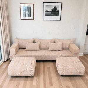 May include: A beige woven sofa with matching ottomans. The sofa has several throw cushions and is placed on a light wood floor. Two framed pictures hang on the wall above the sofa.