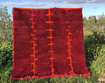 May include: A rectangular, red rug with a textured surface and orange vertical stripe designs. The rug is displayed outdoors against a backdrop of green grass and a blue sky with clouds. The rug's edges are also orange.