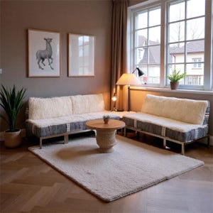 May include: A living room scene with two low sofas, each with cream cushions and grey bases. A round wooden coffee table rests on a large, off-white rug. Two framed pictures adorn the wall, and a floor lamp provides soft lighting.