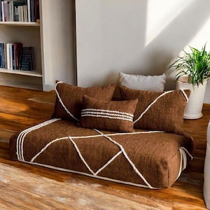 Puede incluir: Un cojín de suelo marrón con costuras geométricas blancas, acompañado de cuatro cojines marrones a juego. Un cojín tiene rayas blancas horizontales. Los cojines están colocados sobre un suelo de madera.