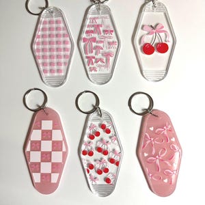 May include: Six clear acrylic keychains with silver key rings. The keychains feature pink and white patterns, including gingham, bows, and cherries. The keychains are in a retro motel style.