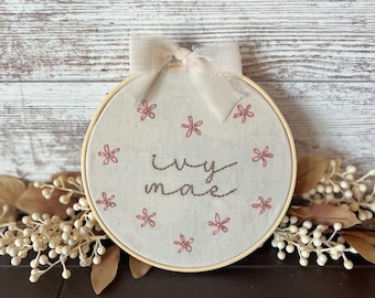May include: A circular wooden embroidery hoop containing off-white fabric with the name "ivy mae" stitched in brown thread. The fabric is decorated with small pink embroidered flowers and topped with a sheer cream-colored ribbon bow.