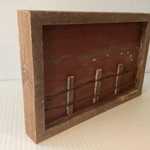 May include: Rustic wooden framed art piece featuring a red background with a barbed wire fence design. The frame is made of weathered wood, adding to the piece's vintage aesthetic. The artwork evokes a rural, country theme.