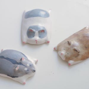 以下が含まれることがあります： 休んでいるげっ歯類の形をした、3つの小さな平らな陶器の動物の置物。1つは暗い縞模様のある灰色、1つは灰色の斑点がある白、もう1つは明るい茶色です。それぞれに小さなピンク色の足先と、シンプルな顔のペイントが施されています。